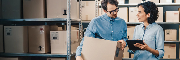 Two people reviewing inventory with a box and tablet in a warehouse.
