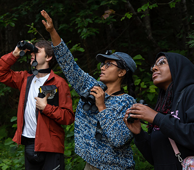 Young woman, with a male and female student on either side of her, holds binoculars in one hand and points toward the tree canopy