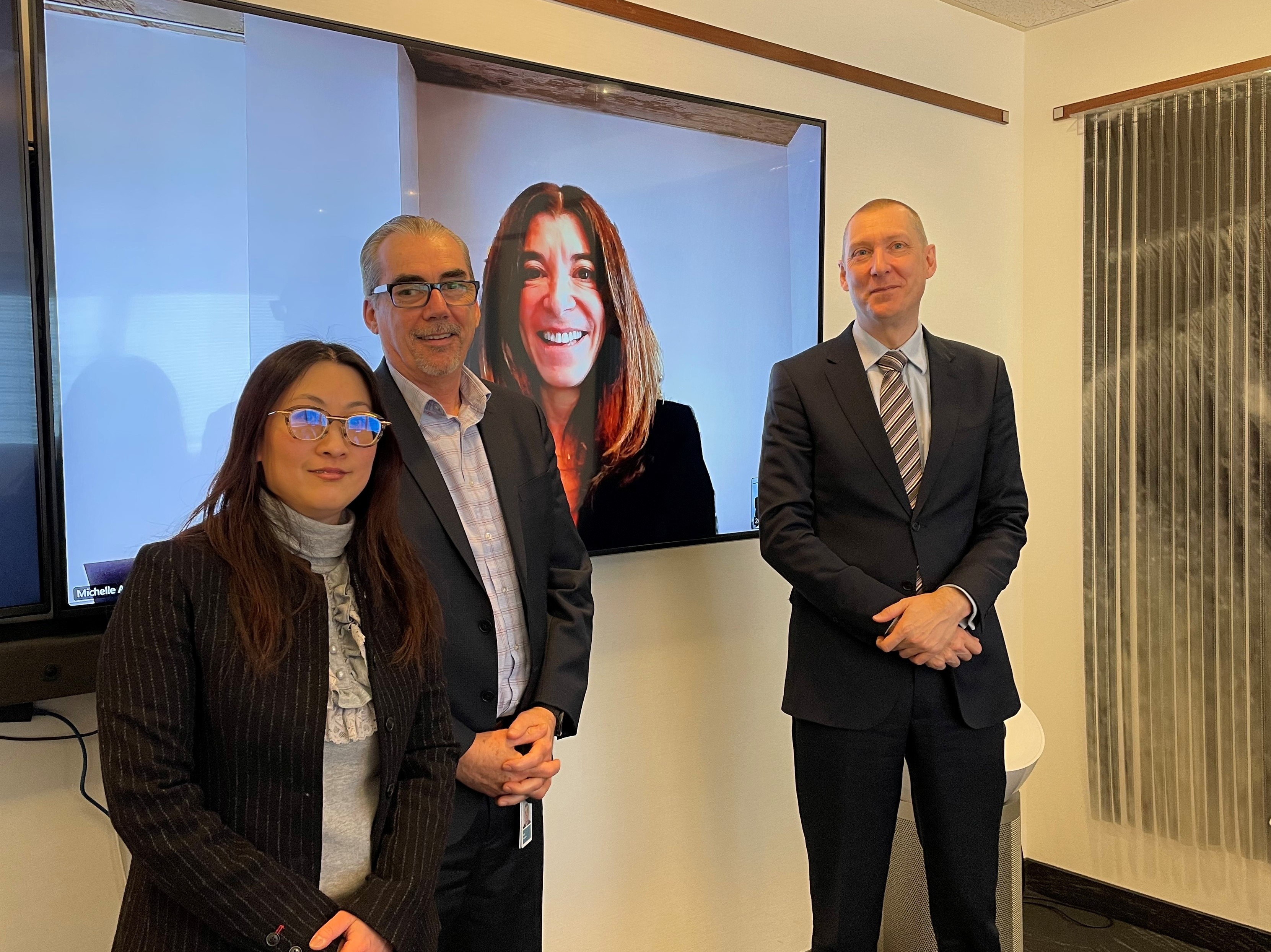 Three people standing in an office beside a large display screen showing a fourth person joining by video. The display shows a person with long hair smiling. In the room, one individual stands on the left wearing glasses and a striped jacket, another stands in the center wearing glasses and a suit, and a third stands on the right in a suit. The office has neutral walls and a mounted screen.