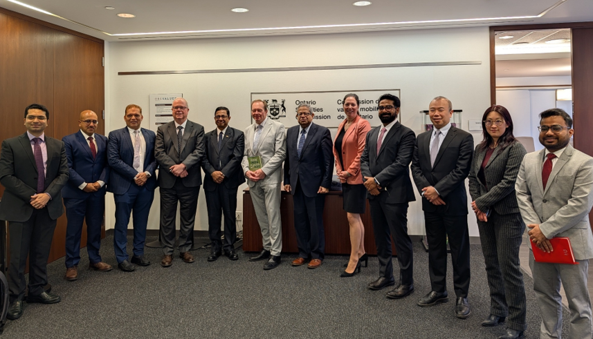 A large group of people dressed in business attire stand in a row inside an office reception area. The group consists of more than a dozen individuals spaced evenly across the room. They are standing in front of a wall displaying a bilingual sign that reads “Ontario Securities Commission” and “Commission des valeurs mobilières de l’Ontario,” along with the OSC logo. The room has carpeted flooring, wood-paneled walls on the left, and a white ceiling with recessed lighting. A small table is positioned against the wall beneath the sign. Everyone is standing still with hands either at their sides or clasped in front.