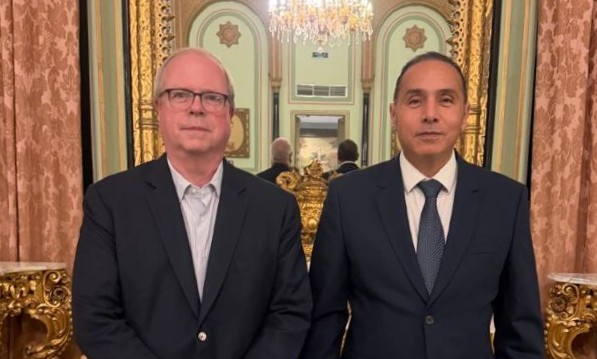 Two individuals in business attire stand side by side in an ornate interior setting featuring gold decorative elements, patterned walls, mirrors, and a chandelier. Both individuals face the camera. The background includes symmetrical architectural details, reflective surfaces, and warm lighting that highlights the intricate decor.
