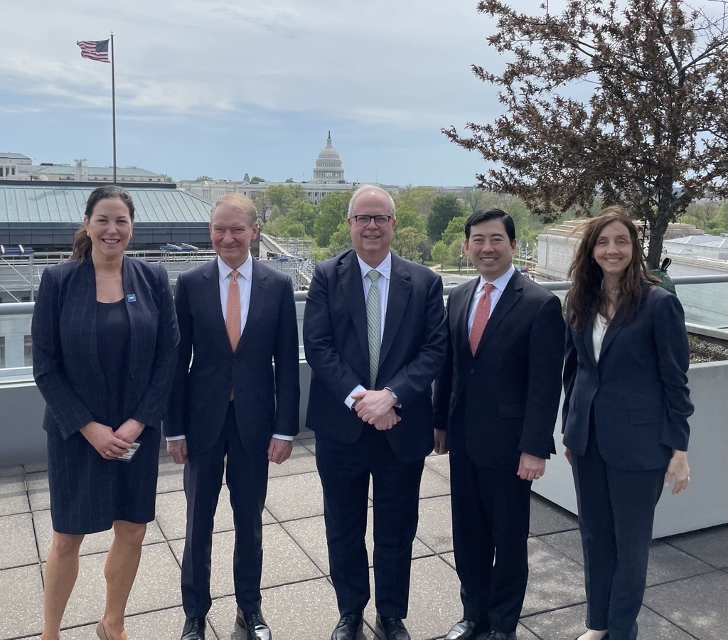 Five people stand side by side outdoors on a terrace or rooftop, wearing formal business attire. The United States Capitol building is visible in the background, along with trees, nearby structures, and an American flag under a lightly overcast sky.