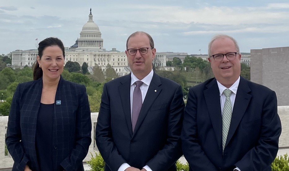 Three people stand side by side outdoors, dressed in formal business attire, with the United States Capitol building visible in the background. Trees and surrounding government buildings are seen under an overcast sky, suggesting a location near Capitol Hill.