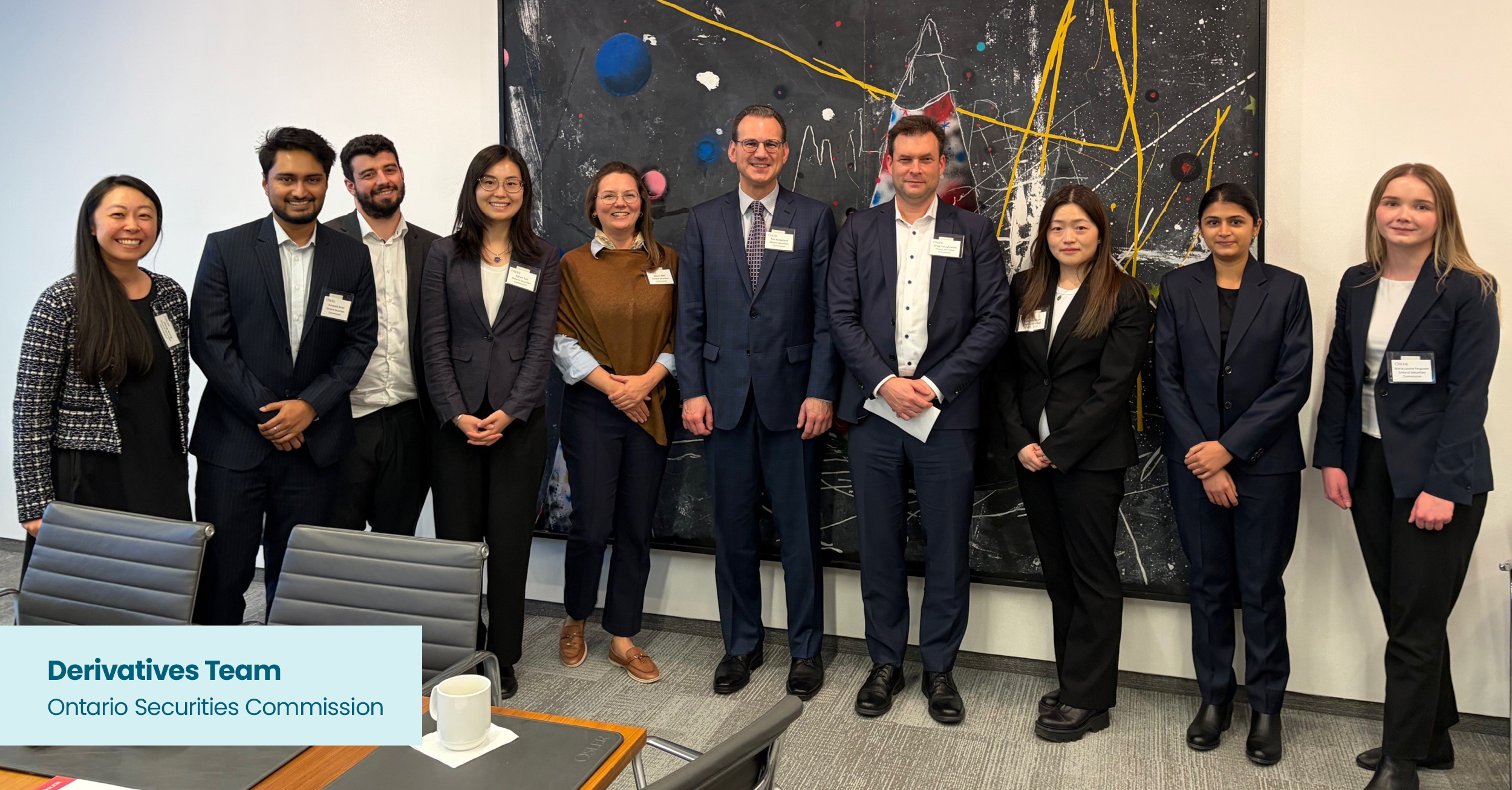 A group of ten people stand side by side in a meeting room, all dressed in professional business attire. They are positioned in front of a large abstract painting featuring black, yellow, white, red, and blue elements. A conference table with chairs, a white mug, and meeting materials is in the foreground. A small graphic label in the lower left corner reads “Derivatives Team, Ontario Securities Commission.