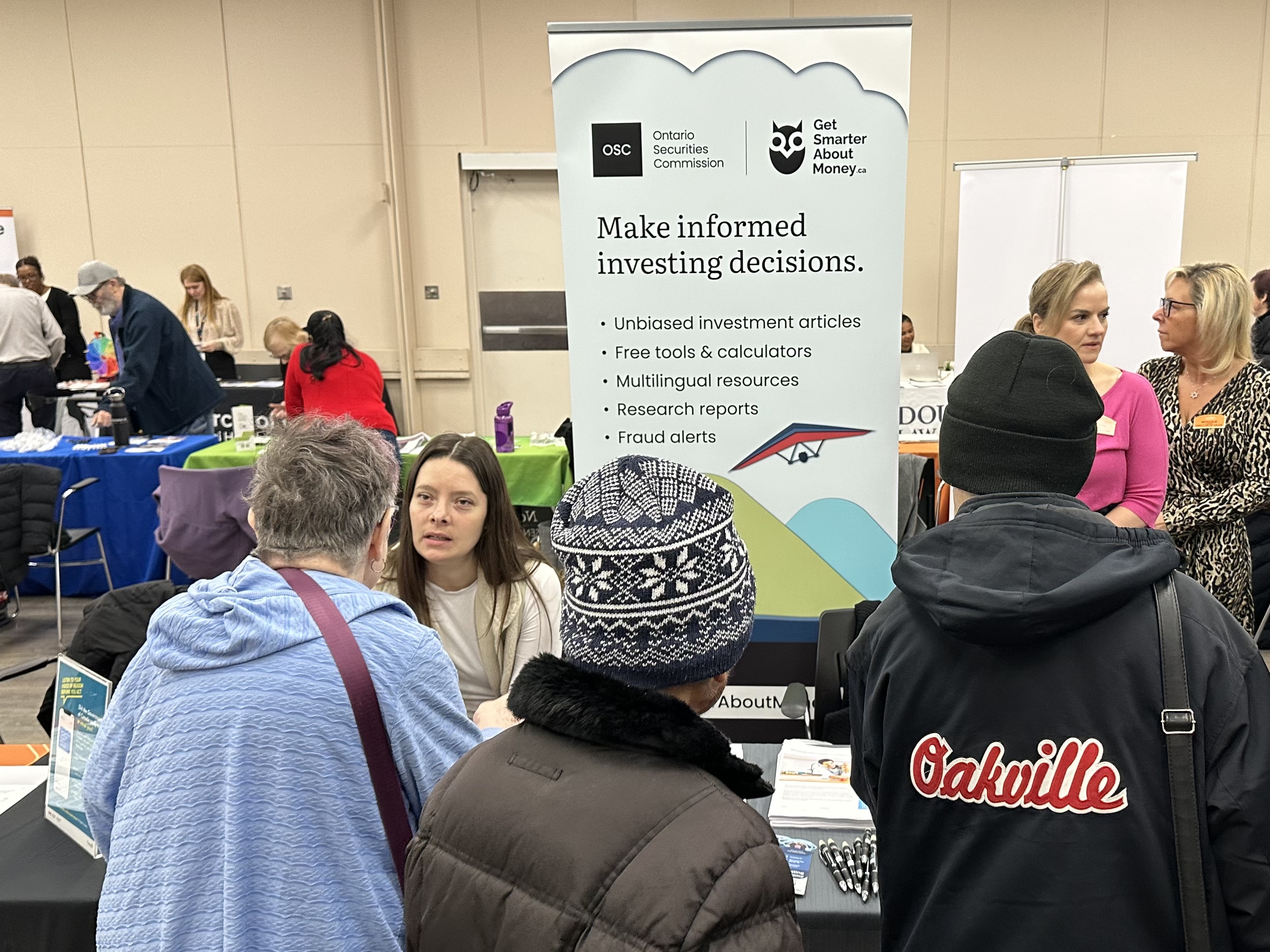A public event or information fair with people gathered around a booth featuring an Ontario Securities Commission and Get Smarter About Money banner. The banner lists resources for making informed investing decisions. Tables with brochures and displays are visible, and several attendees stand in front of the booth.