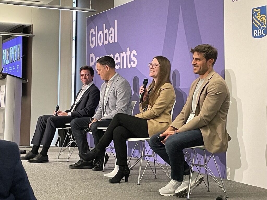 Four individuals sit on stage during a panel discussion at a conference event. Each individual is seated on a chair and holding a handheld microphone. A purple backdrop behind the panel displays the words “Global Payments,” with an RBC logo visible in the upper right corner. One individual is speaking into a microphone while the others face forward. A large screen on the left shows participant images or presentation content. The setting appears to be a modern conference room with bright lighting and a small audience area visible in the foreground.