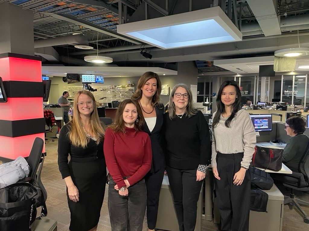 A group of five people standing together in a newsroom environment. They are positioned in front of desks, monitors, and office equipment. The room has overhead lighting, exposed ceiling fixtures, and several employees working at stations in the background.