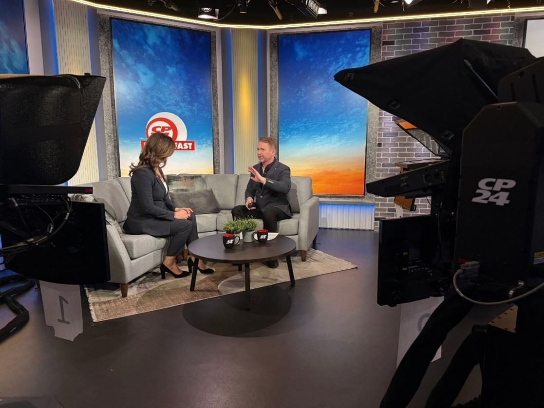 A television studio set with two people seated on a grey couch, engaged in an interview. Large studio cameras and equipment frame the foreground. Behind the couch are two tall screens displaying gradient blue‑to‑orange backgrounds and a CP24 Breakfast logo. A small round coffee table with mugs and a plant sits between the two guests.