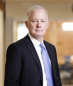 A professional portrait of a person wearing a dark business suit and a light‑colored tie, standing indoors in a softly lit office or institutional setting. The subject is positioned slightly off‑center and faces the camera, with a shallow depth of field that blurs the background of warm-toned interior architecture and ambient lighting.