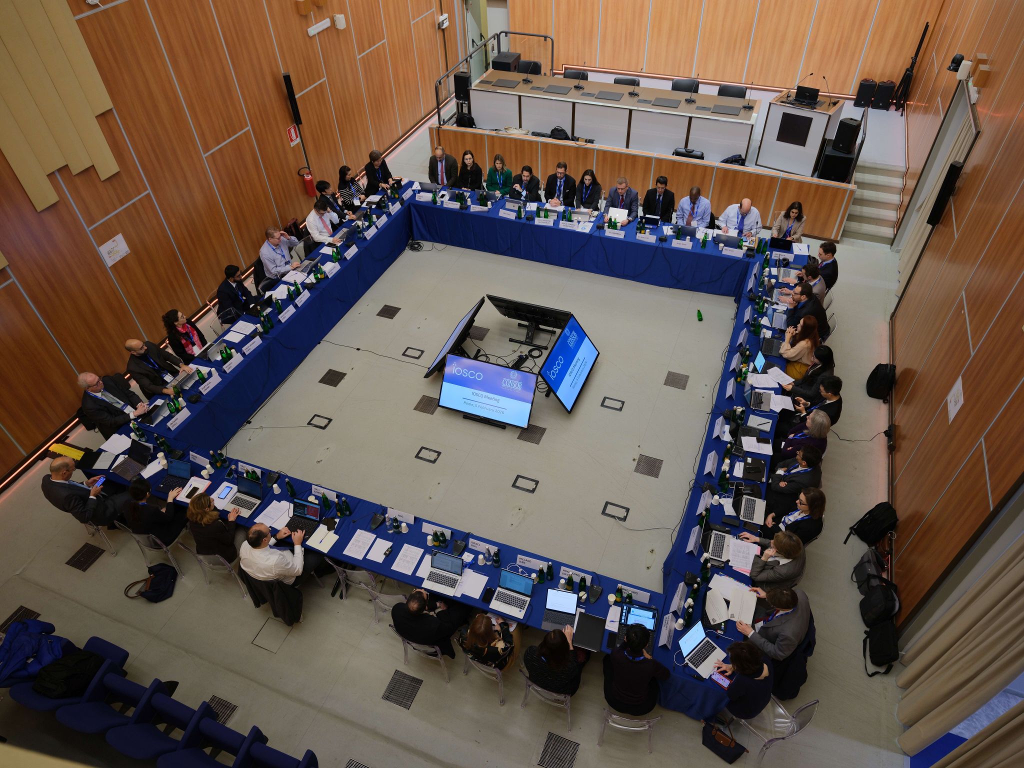 Wide overhead photo of a large square meeting table with delegates seated around all four sides. Screens in the centre display IOSCO materials, and participants have laptops, documents, and microphones in front of them