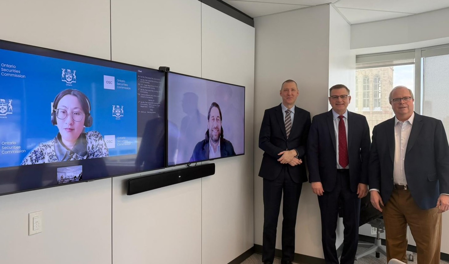 An office setting with two large wall‑mounted screens showing a video meeting. One screen displays a person wearing a headset with an Ontario Securities Commission background, and the second screen displays another participant. Three people stand in the room next to the screens, all wearing business attire, with windows showing buildings outside.
