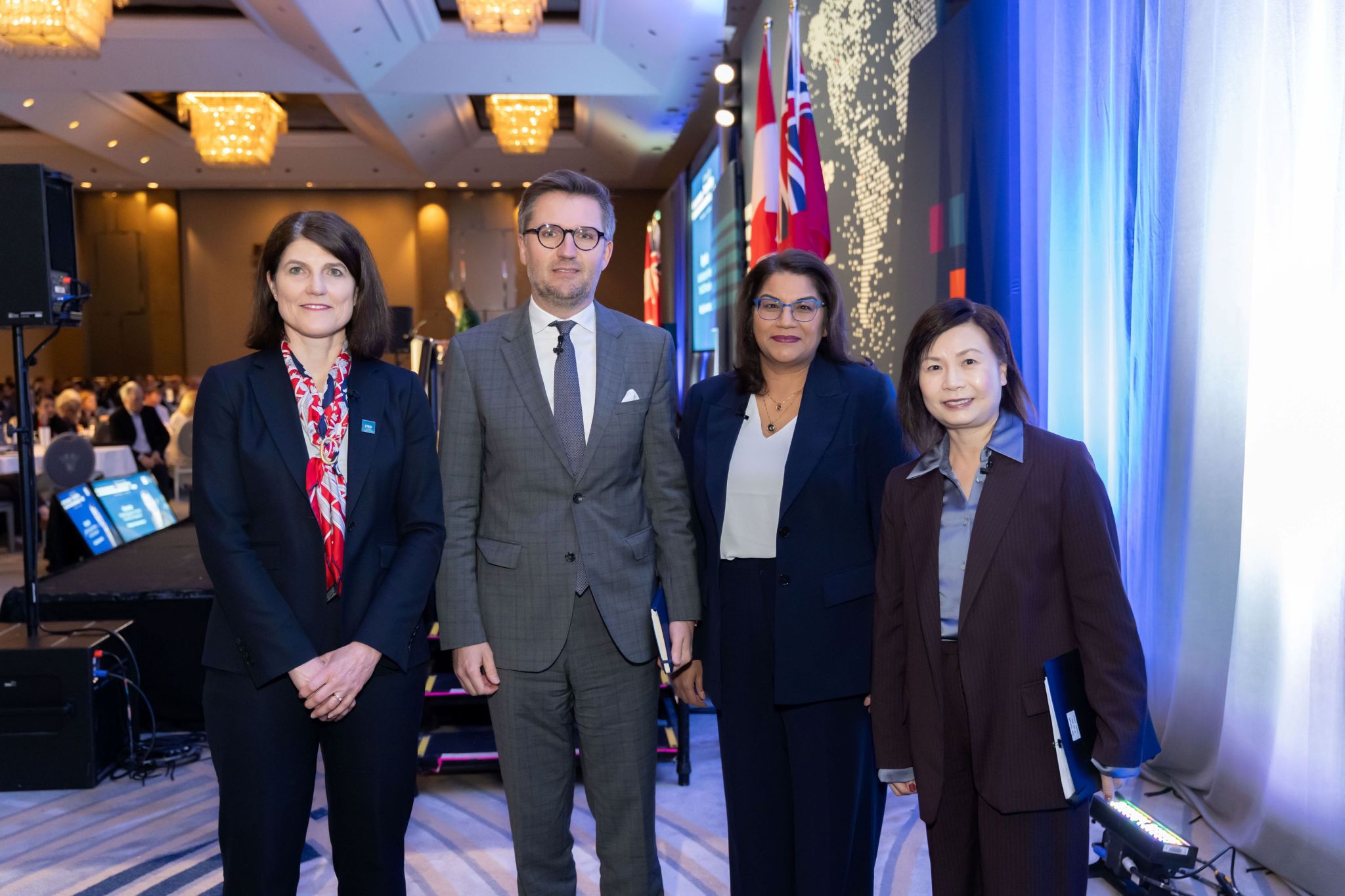 Four conference participants stand in a line near a stage inside a large ballroom, wearing business attire and conference microphones. Behind them are OSC Dialogue–branded graphics, a large digital screen, Canadian and Ontario flags, stage lighting, and audiovisual equipment, with seated attendees visible in the background under chandelier lighting.