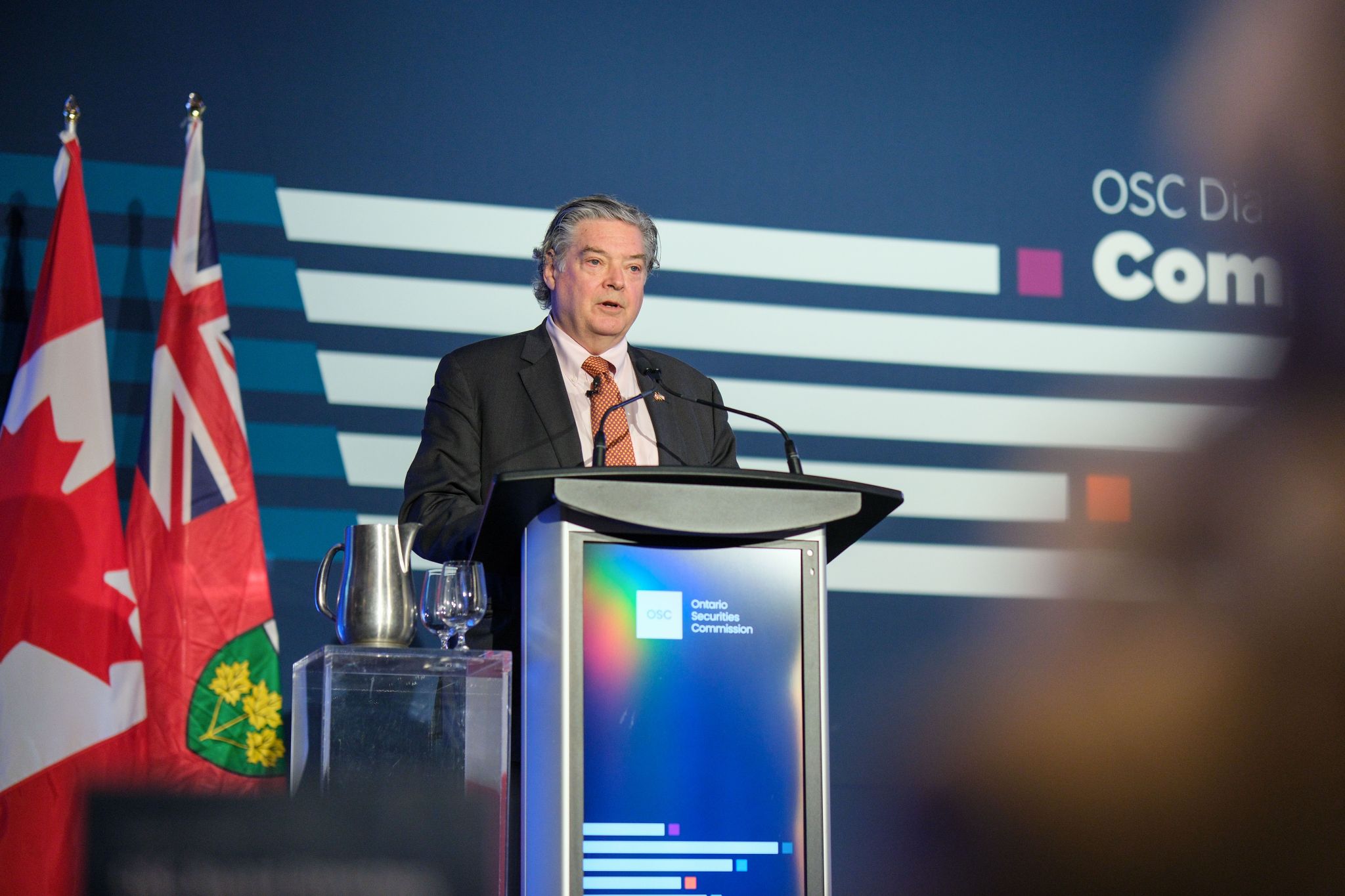 A speaker stands at a podium branded with the Ontario Securities Commission logo, addressing an audience on a stage with a blue graphic backdrop reading “OSC Dialogue” and partial conference graphics. The Canadian and Ontario flags are positioned behind the podium, and a water pitcher and glass sit on a clear side stand nearby.