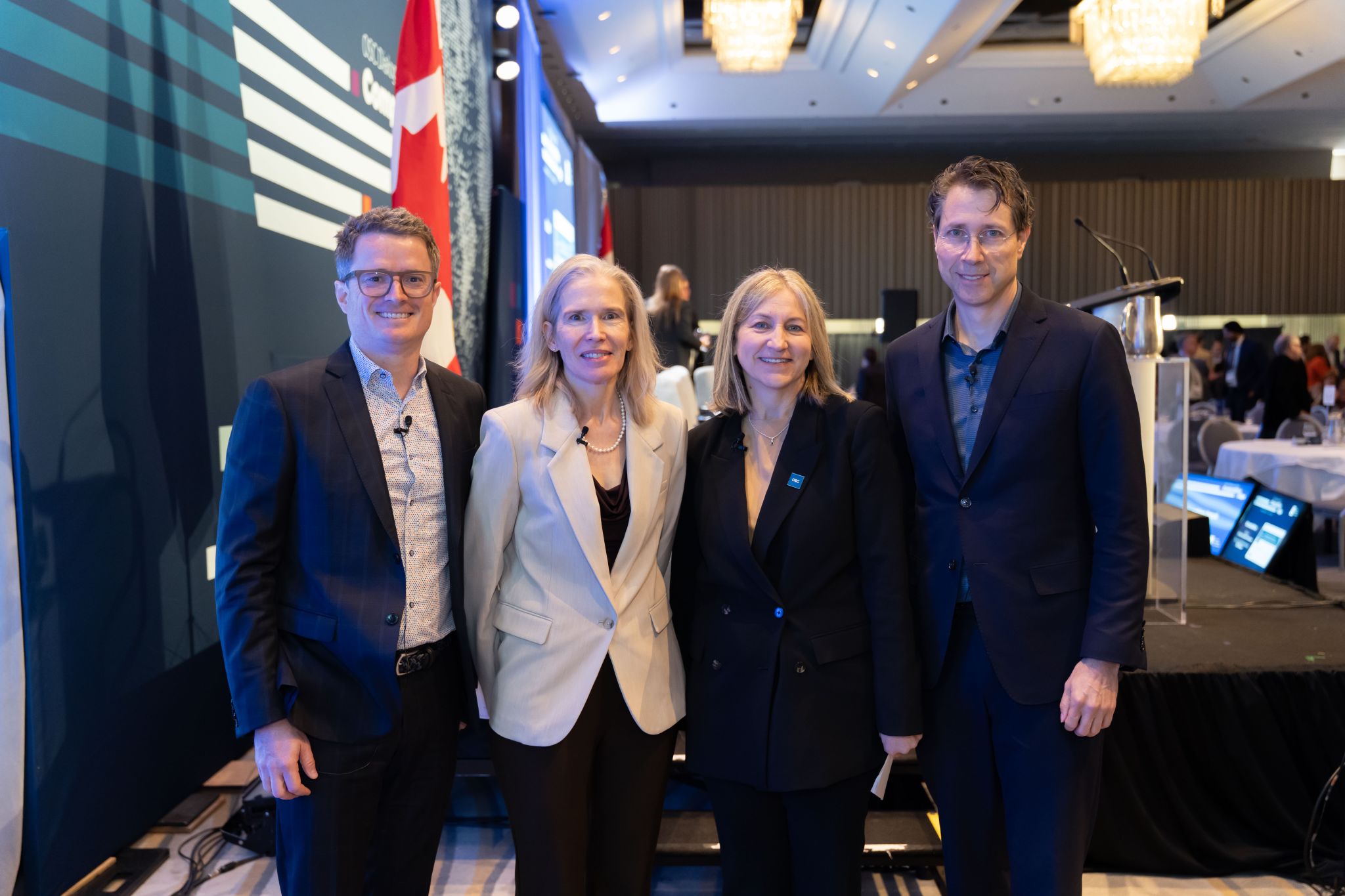 Four conference speakers stand side by side on a stage area in a ballroom, wearing business attire and lapel microphones. Behind them are a large OSC Dialogue-branded screen, a Canadian flag, and stage equipment, with banquet tables, lighting, and attendees visible in the background.