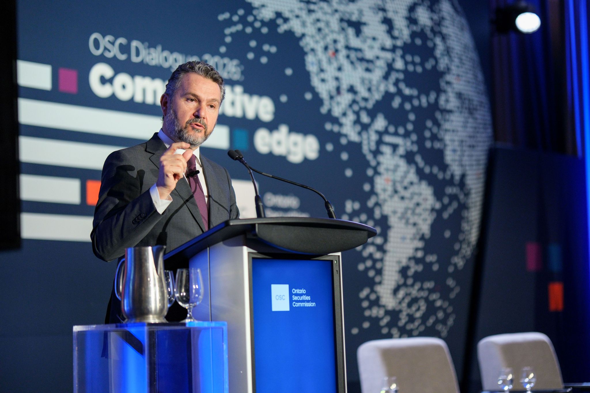 A conference speaker stands at a podium branded with the Ontario Securities Commission logo, addressing an audience on stage. Behind the podium is an OSC Dialogue backdrop with the words “Competitive edge” and a stylized world map graphic, with stage lighting, microphones, and a water pitcher and glass on a nearby stand.
