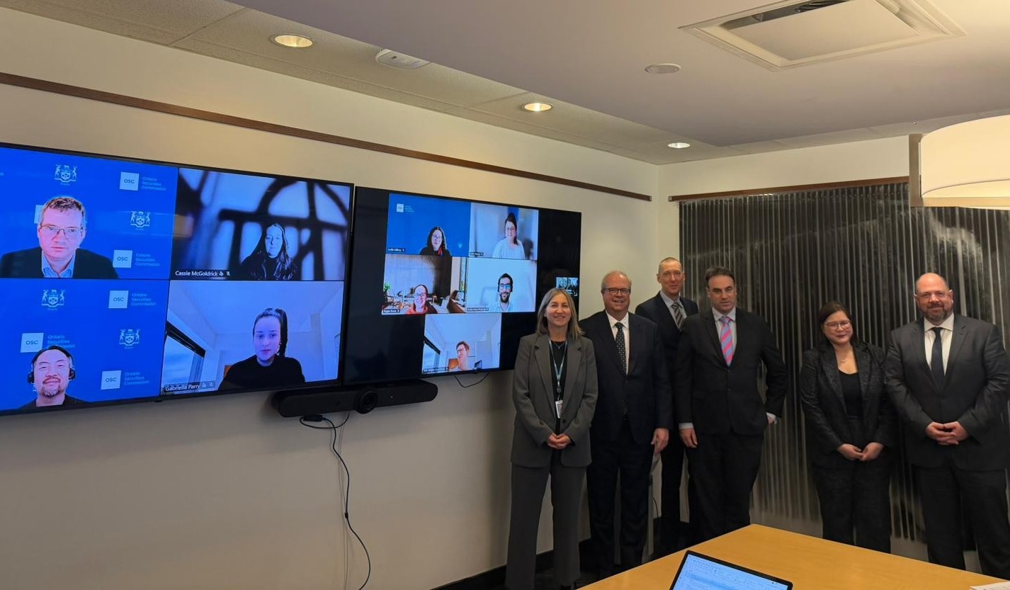 A conference room with two large screens displaying multiple participants in a virtual meeting. Several people are standing in a row in front of the screens, all wearing business attire. The room has overhead lighting, neutral walls, and a wooden table in the foreground.