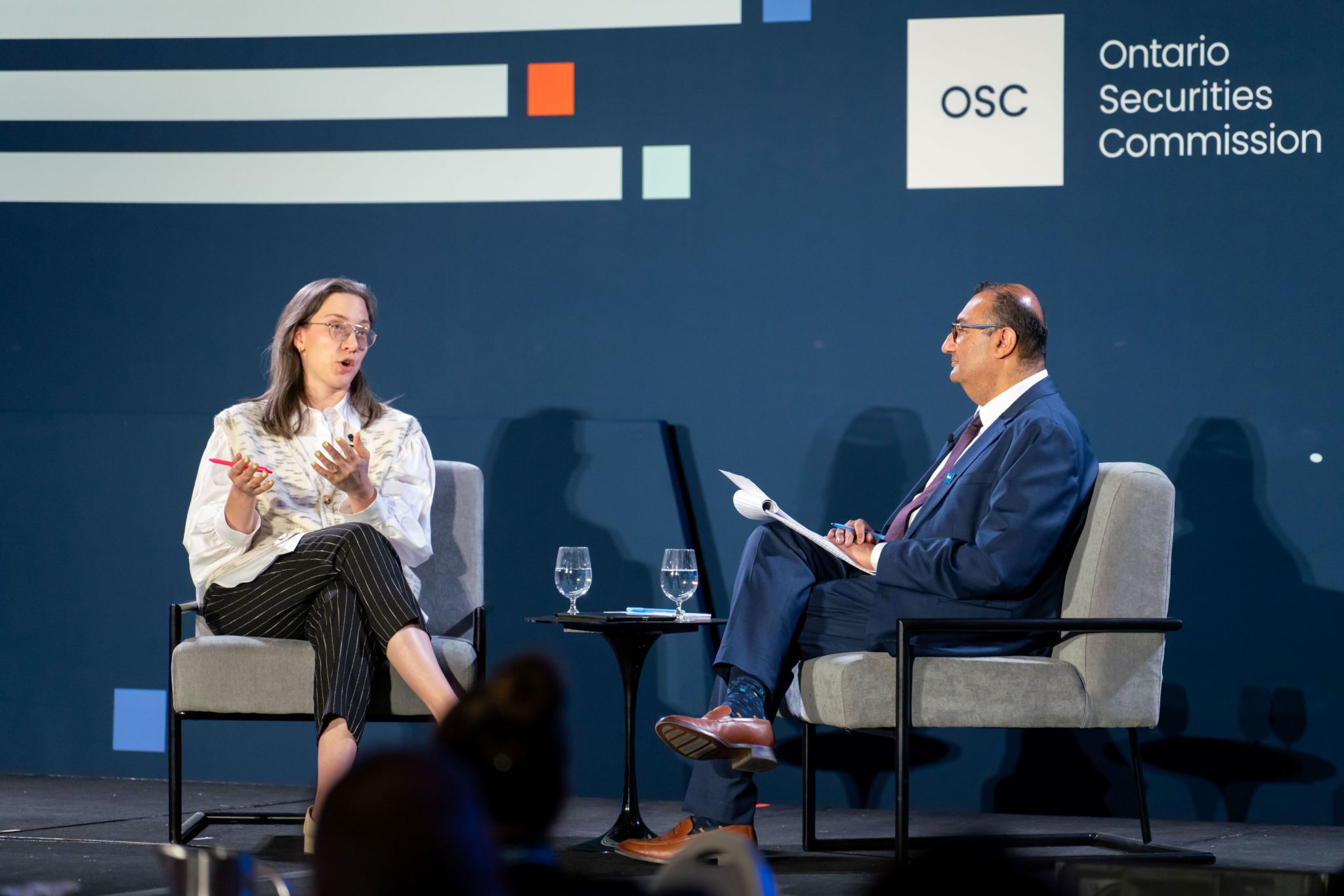 Two speakers sit in armchairs on a stage during a moderated conversation at an OSC Dialogue event. One speaker gestures while speaking, and the other listens while holding papers. Between them is a small table with two water glasses. Behind them is an OSC Dialogue–branded backdrop displaying the Ontario Securities Commission logo and graphic lines.