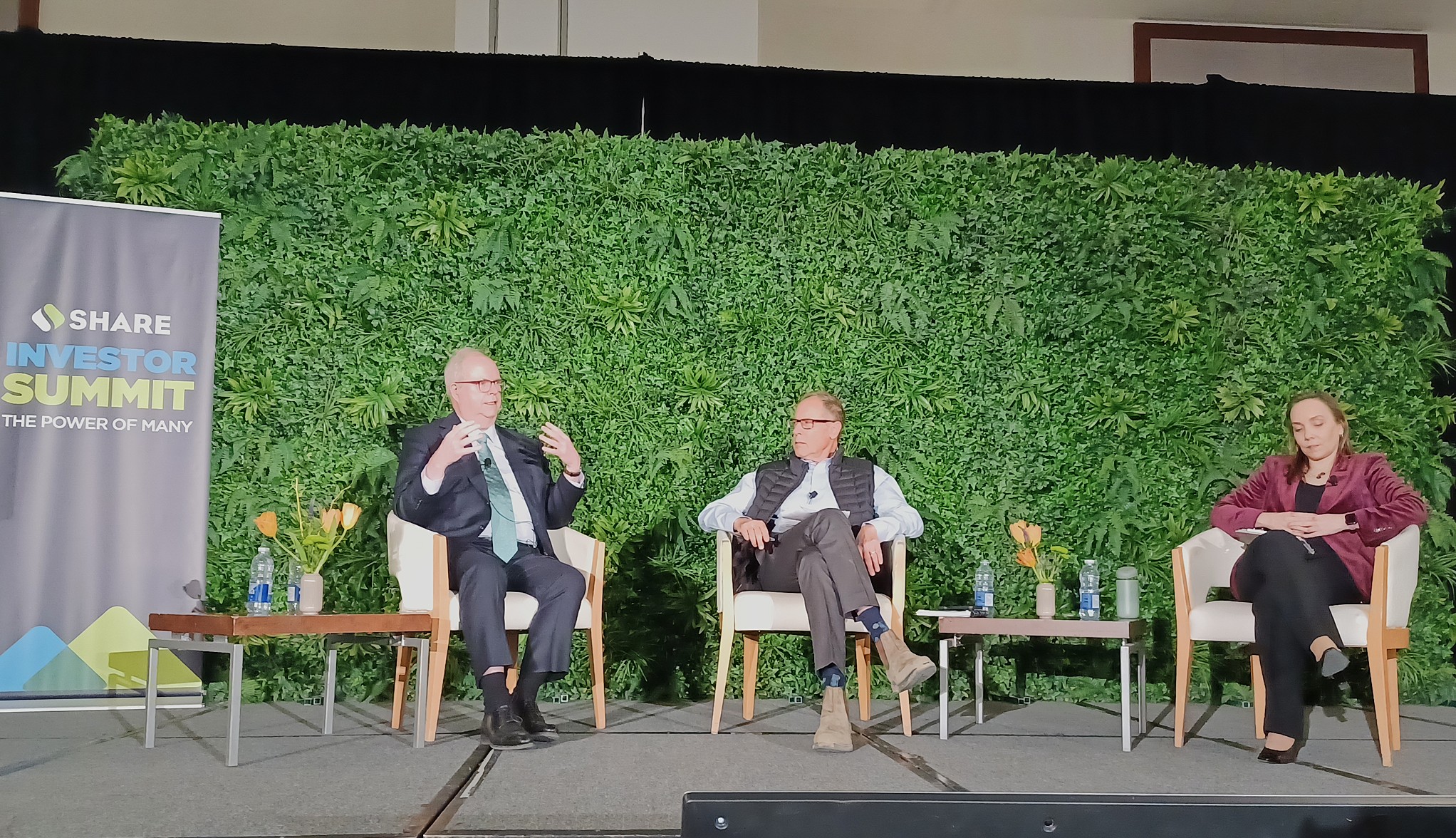Three panelists are seated on a stage during a conference discussion, each in an armchair with small side tables holding water bottles and floral arrangements. A large green plant wall forms the backdrop, and a standing banner on the left reads “SHARE Investor Summit: The Power of Many.” One panelist gestures while speaking, while the other two listen. The stage is softly lit, with the audience out of frame, creating a formal conference setting.
