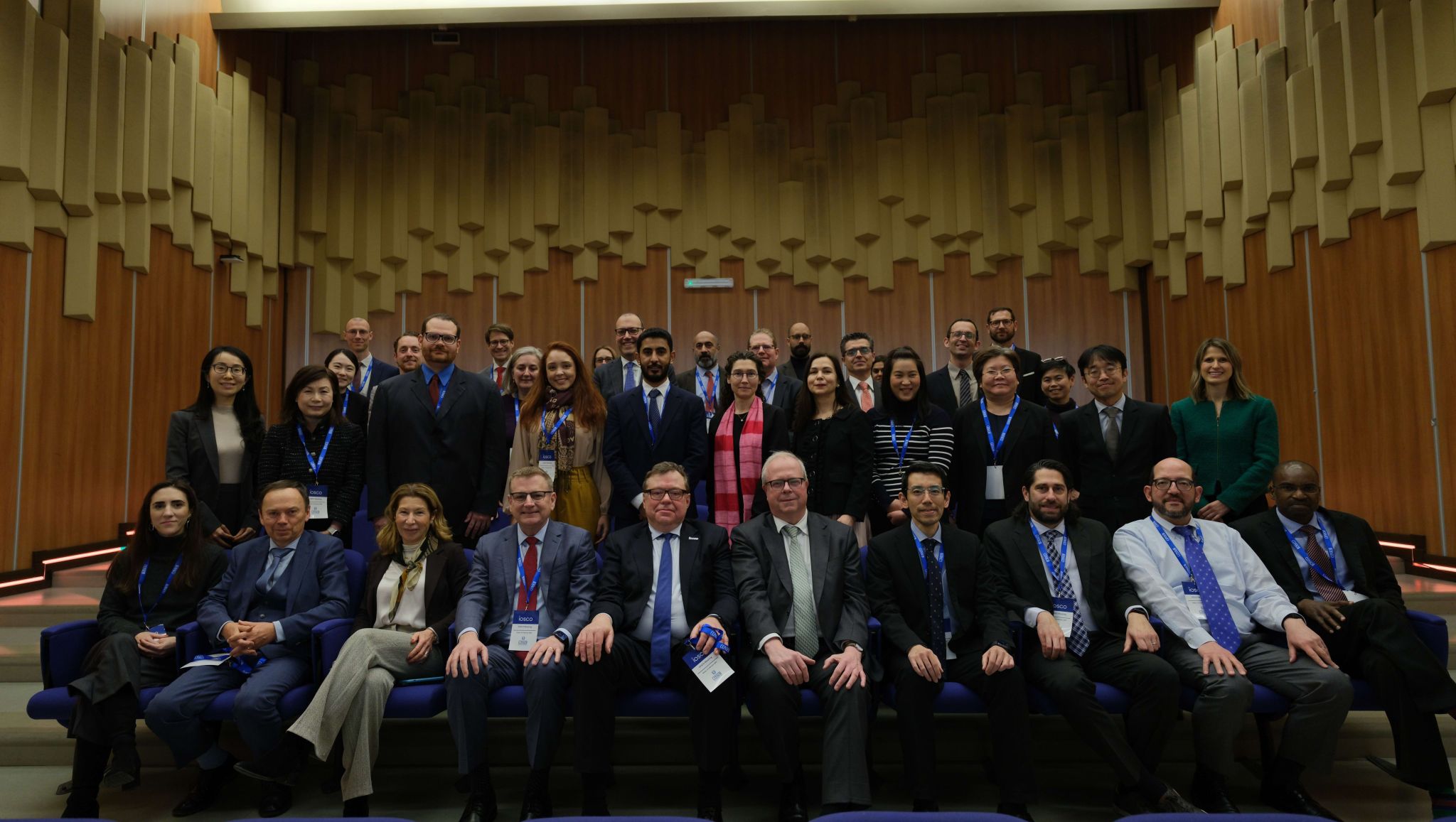 Group photo of approximately 30 conference participants posed in several rows inside a large auditorium with wood-panelled walls and tiered seating. Attendees are dressed in business attire, many wearing conference lanyards, and are gathered in the centre of the room for the photo.