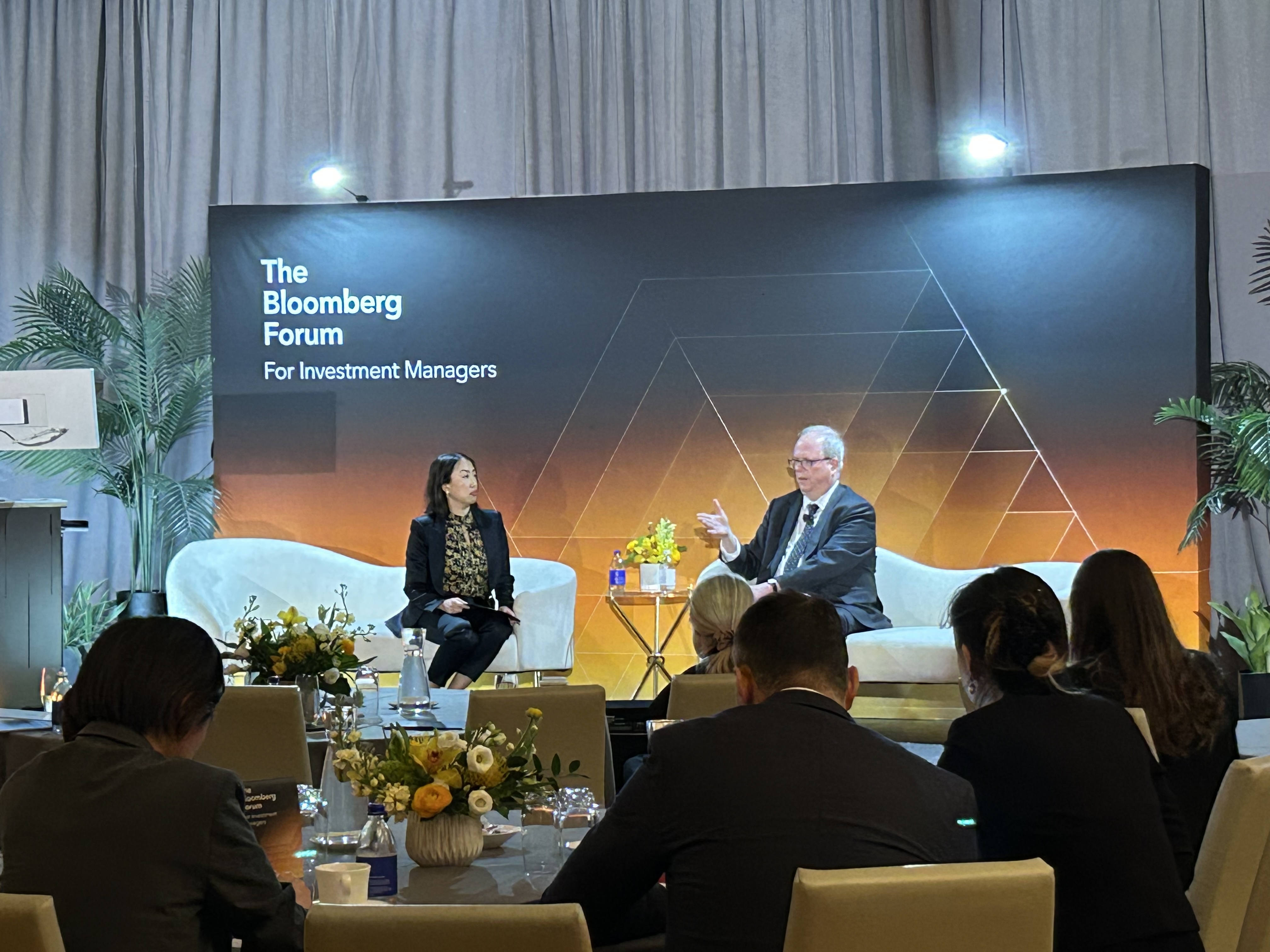 A moderated discussion takes place on stage at The Bloomberg Forum for Investment Managers. Two speakers are seated on white armchairs facing an audience seated at round tables. A branded backdrop behind the stage reads “The Bloomberg Forum for Investment Managers” with a geometric line design in dark and warm gradient tones. Between the speakers is a small side table holding water bottles and a floral arrangement. In the foreground, audience members are seen from behind, seated at tables with glasses, notebooks, and tabletop floral centerpieces, in a softly lit conference setting.