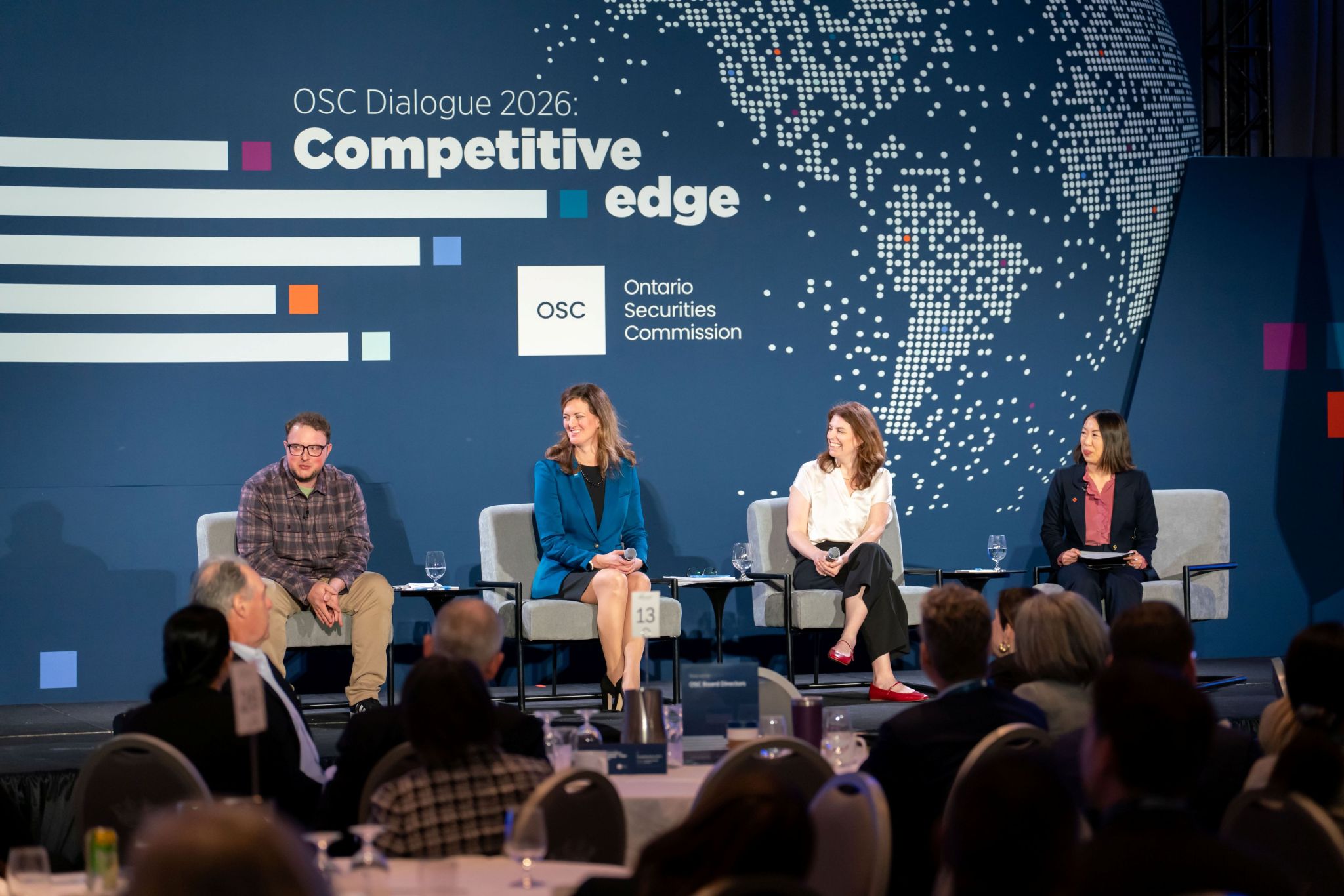 Four panelists are seated on stage in armchairs during an OSC Dialogue 2026 panel discussion, facing an audience seated at round tables in a ballroom. The stage backdrop reads “OSC Dialogue 2026: Competitive edge” and features the Ontario Securities Commission logo and a dotted world map graphic. Each panelist has a microphone, with small side tables holding water glasses, and the audience is visible in the foreground.