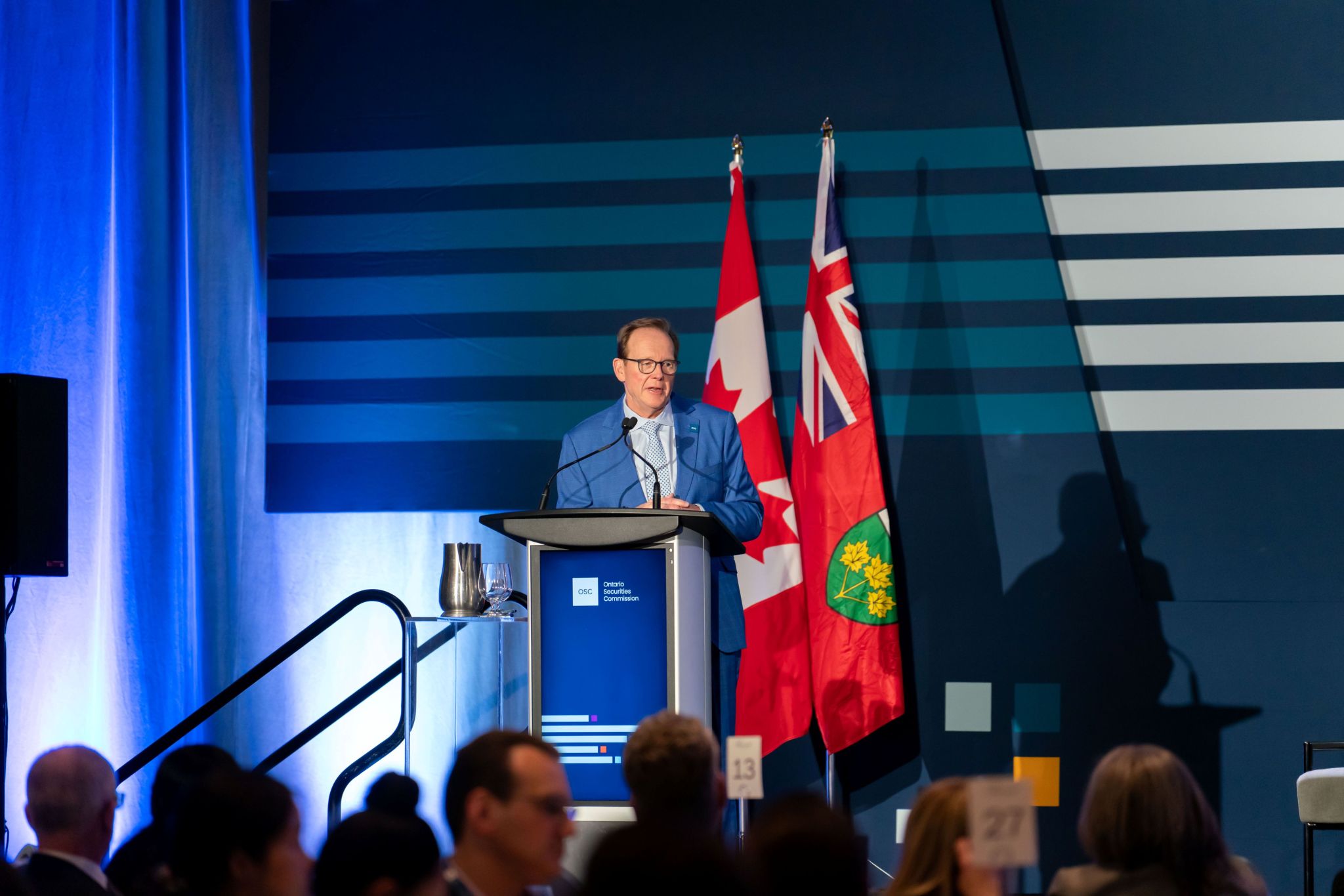 A speaker stands at a podium branded with the Ontario Securities Commission logo, addressing an audience in a large ballroom. Behind the podium are the Canadian and Ontario flags, OSC Dialogue–style graphic stripes on a blue backdrop, and stage lighting. A water pitcher and glass sit on a small stand beside the podium, with audience members visible in the foreground.