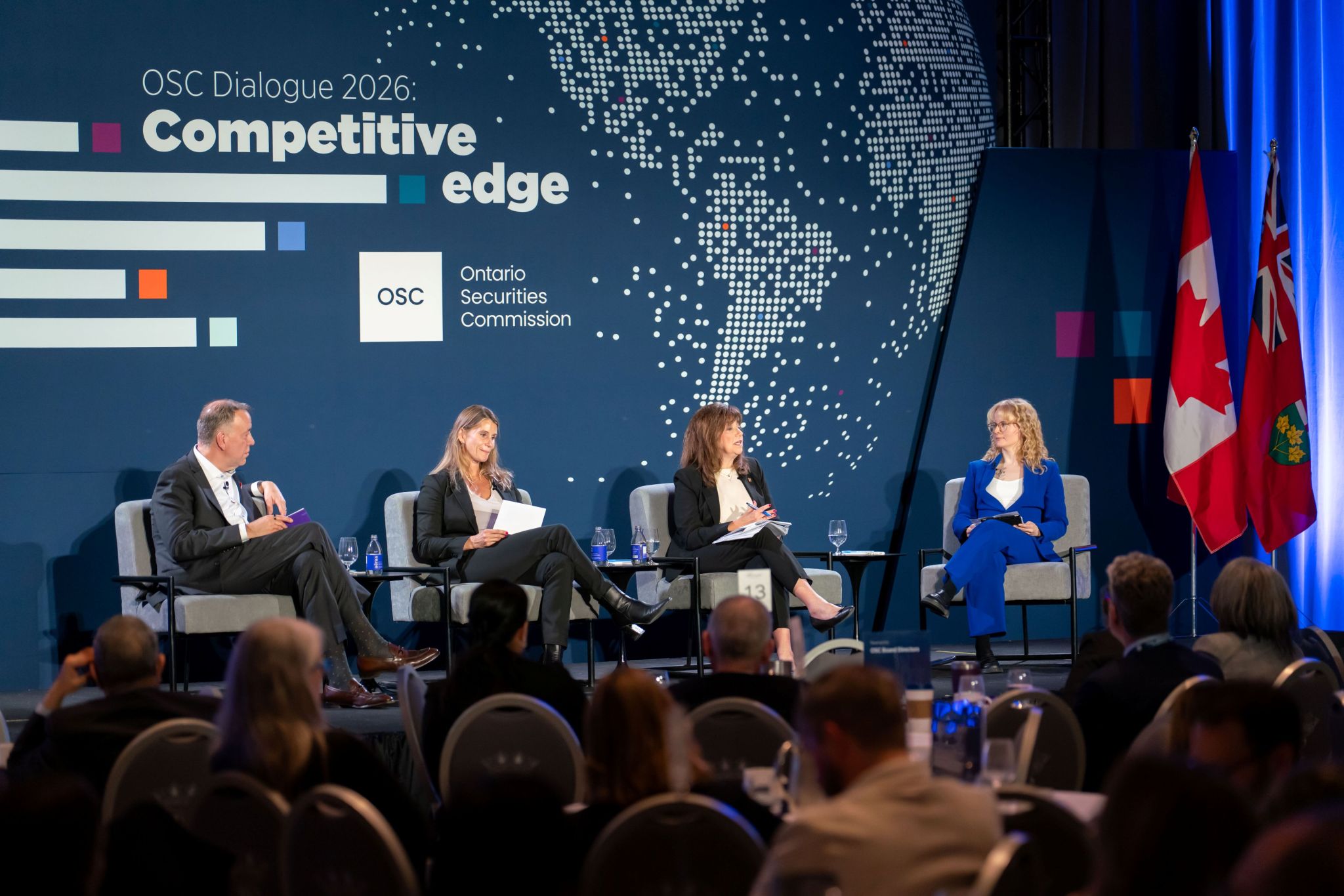 Four panelists are seated in armchairs on a stage during an OSC Dialogue 2026 panel discussion, facing an audience seated at round tables. The backdrop reads “OSC Dialogue 2026: Competitive edge” and features the Ontario Securities Commission logo and a dotted world map graphic. Small side tables with water bottles and glasses sit beside the panelists, with Canadian and Ontario flags visible at the side of the stage.