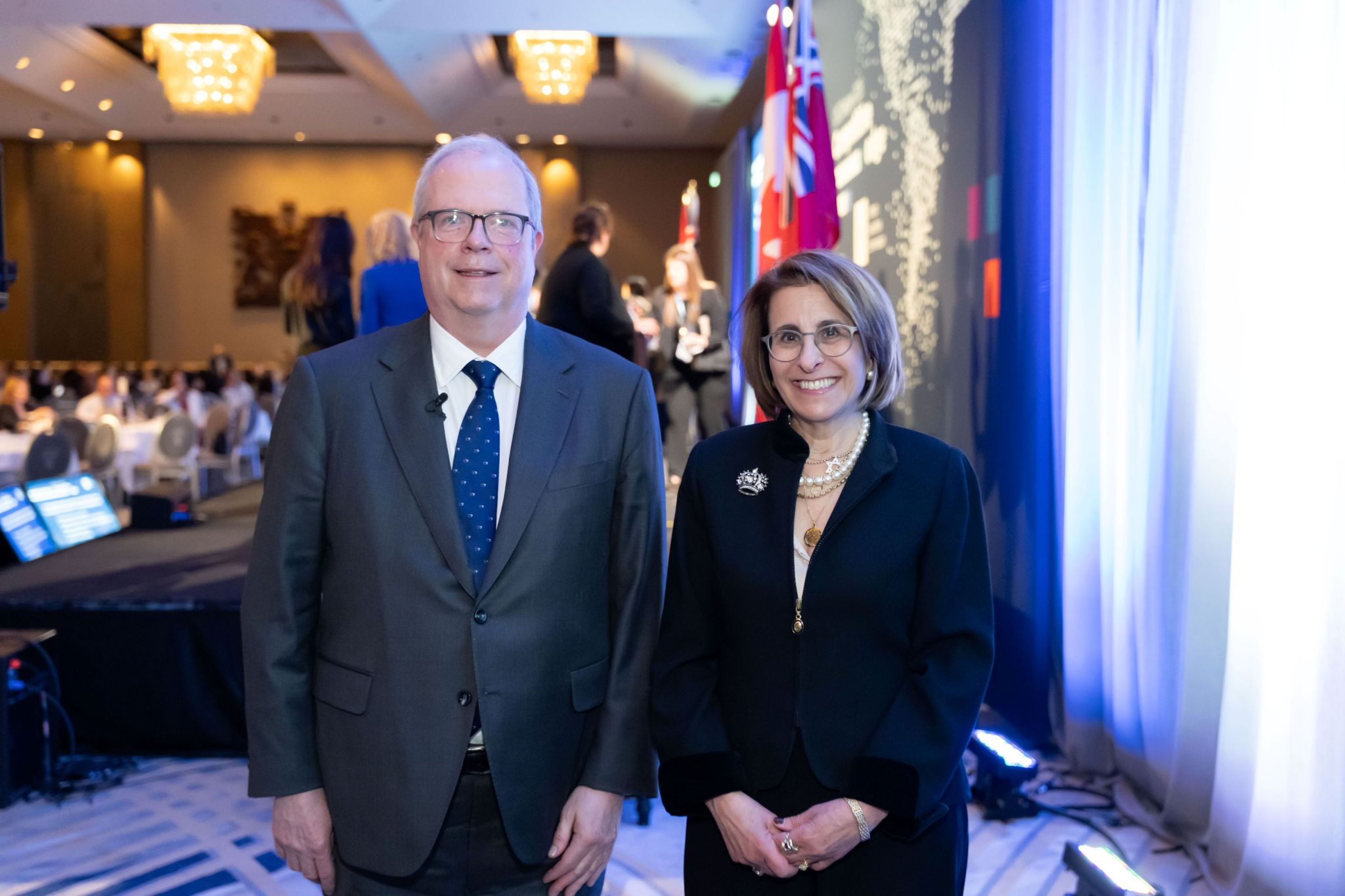 Two conference attendees stand side by side near the stage area of a large ballroom, wearing formal business attire. Behind them are OSC Dialogue–branded stage graphics, Canadian and Ontario flags, stage lighting, and audiovisual equipment, with other attendees and banquet tables visible in the background under chandelier lighting.