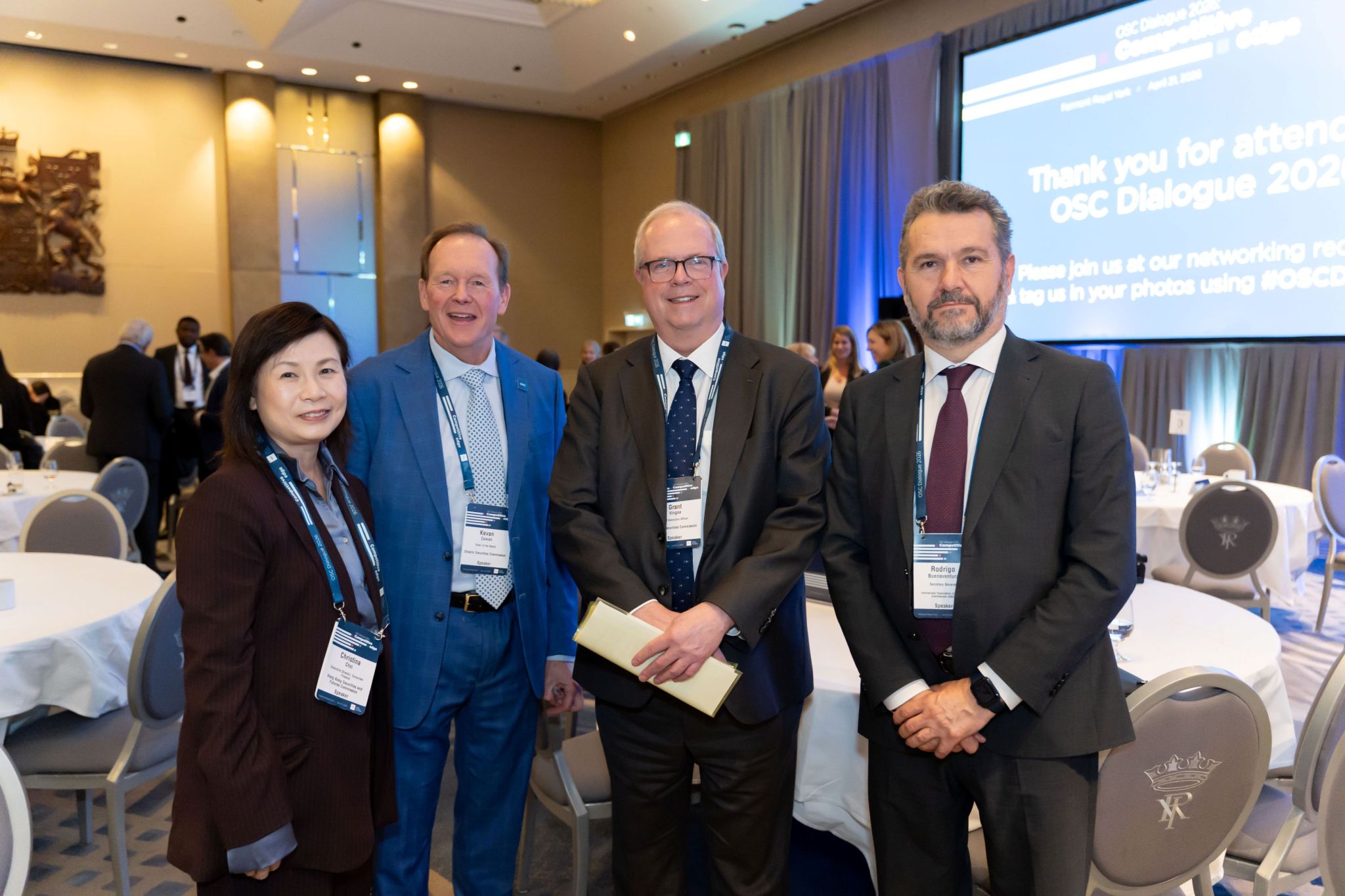Four conference attendees wearing name badges stand together in a hotel ballroom set with round tables and chairs. Behind them, a large presentation screen reads “Thank you for attending OSC Dialogue 2026,” with other attendees and stage lighting visible in the background.