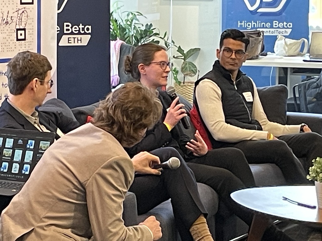 A small group discussion is taking place in a modern office setting. Several individuals are seated on a couch and chairs arranged in a semicircle. One person holds a handheld microphone while speaking, and another individual seated nearby also holds a microphone. Branded signage is visible in the background, including banners that read “Highline Beta” and “ETH.” The setting includes indoor plants, glass walls, and office furniture, contributing to a collaborative event atmosphere.
