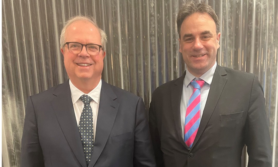 Two people standing side‑by‑side in front of a vertically striped metallic wall. Both individuals are wearing dark business suits. The person on the left has short light hair, glasses, and a patterned tie. The person on the right has short dark hair and a striped tie with pink and blue diagonal bands. Both are facing the camera.